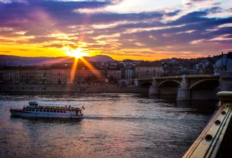 A Danube river cruise ship sailing at sunset with city lights visible on the horizon